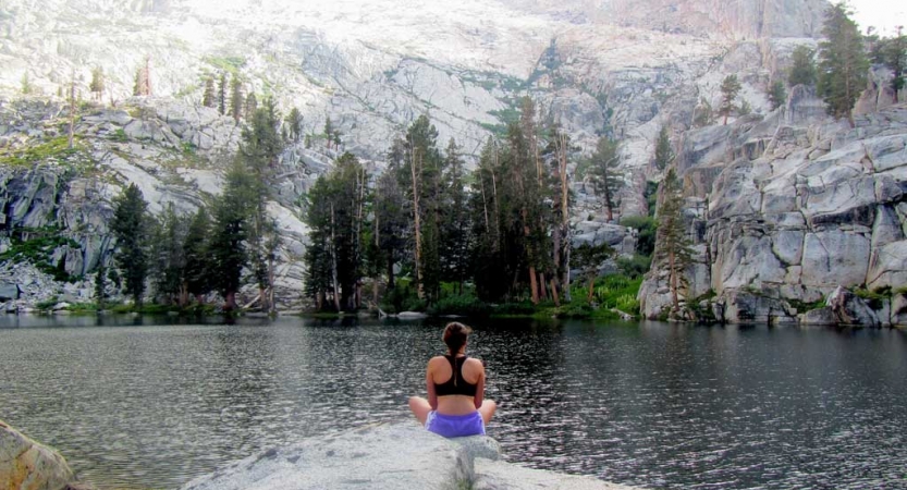 A person faces away from the camera and looks out at an alpine lake. The lake is framed by a steep rocky shore dotted with evergreen trees. 
